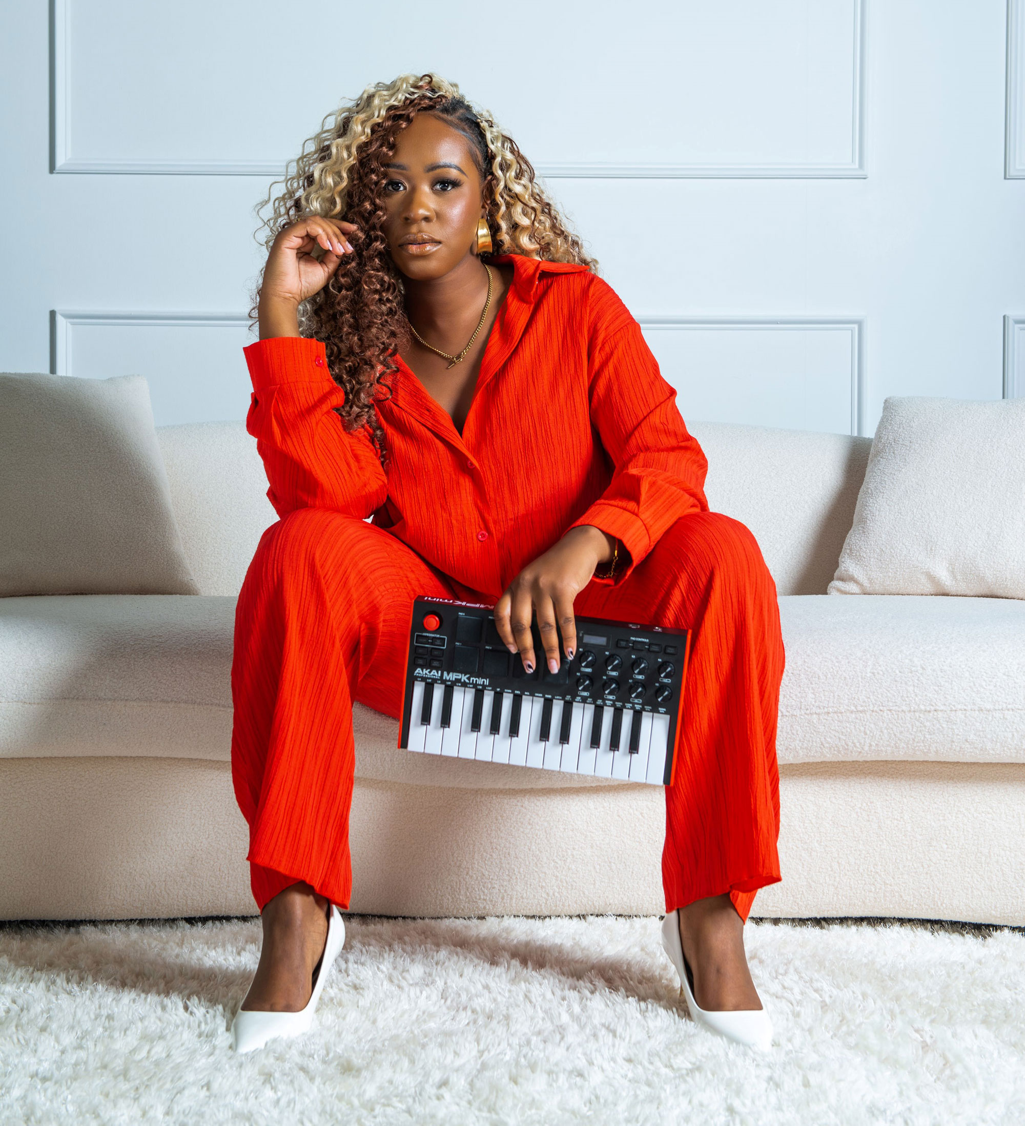 Desarae Dee sitting on a white couch in a red dress suit with white heels on a fluffy white carpet. Desarae is holding a small keyboard in her hands and looking at the camera . She has blond and brown curly hair and is wearing wide gold earrings. 