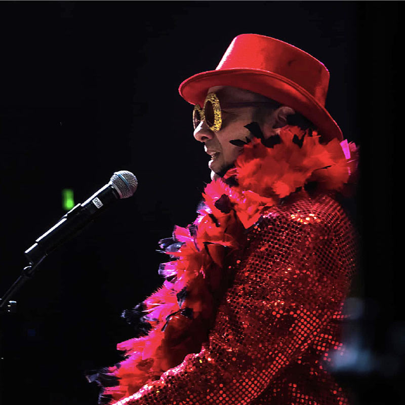 A photo of Captain Fantastic seated at a piano singing into a microphone, wearing a sparkly red jacket, red feather boa, red top hat, and gold sparkly sun glasses.